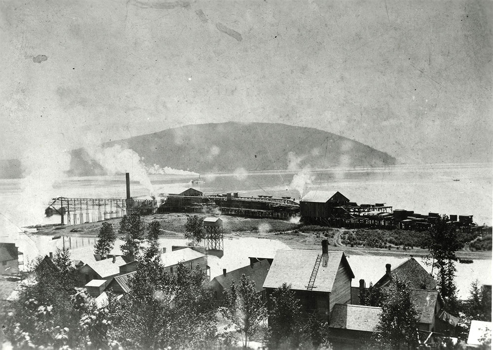 Photographie en noir et blanc d’une ville au bord de l’eau. On peut y repérer des arbres et des maisons au premier plan. Derrière, une scierie se trouve au bord de l’eau et de la fumée s’en échappe. Au loin, un bateau à vapeur avance sur l’eau, avec une montagne en arrière-plan.