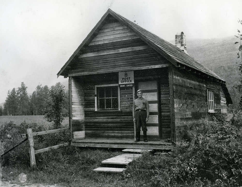 Photographie en noir et blanc d’un homme sur le porche d’un bâtiment en bois rond sur lequel on peut lire « Post Office Mount Cartier (Bureau de poste Mount Cartier) ». Le bâtiment est entouré de buissons et d’arbres, avec une petite clôture en bois à la gauche. Des années plus tard, James Ozero a construit un bureau de poste sur son propre terrain, plus près de la gare.