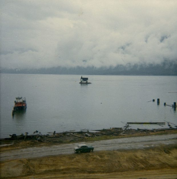 Photographie couleur d'une petite église au loin, entourée d'une grande étendue d'eau. Un bateau est dans l’eau à gauche et des pieux de quai se trouvent à droite. Sur la rive, une voiture roule sur une route parallèle à la rivière. Des grumes et des billots de bois sont éparpillés sur le rivage. Des nuages bas couvrent les montagnes en arrière-plan.