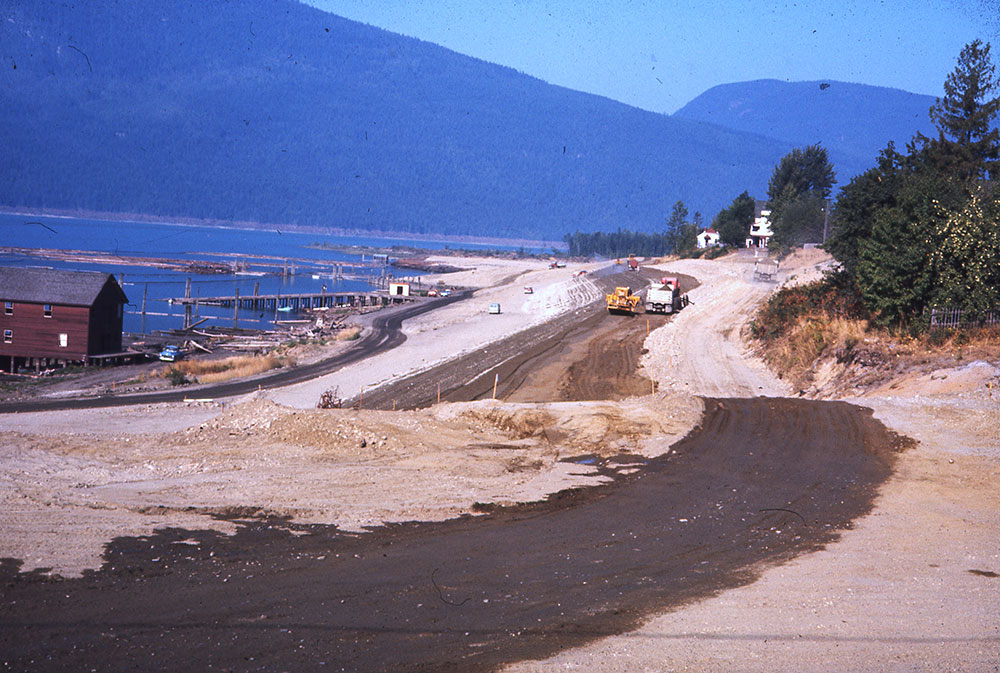 Photographie couleur montrant un terrain en train d'être nivelé et dégagé au bord de l'eau. On aperçoit de gros camions de construction et des voitures au loin. Un bâtiment se trouve au bord de l'eau à gauche. Il y a des montagnes en arrière-plan et des arbres à droite.