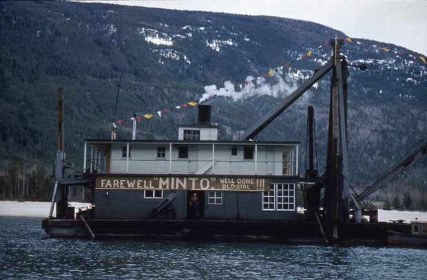 Une drague sur l’eau affiche un panneau en bois sur lequel on peut lire : « Farewell “MINTO”. Well done old girl!!! (Adieu “MINTO”. Bien joué ma vieille!!!) ». Un homme se tient dans l’embrasure de la porte, sous le panneau. Une guirlande de fanions triangulaires colorés est attachée de la cheminée jusqu’à l’avant du bateau. Une montagne se trouve à l’arrière-plan.