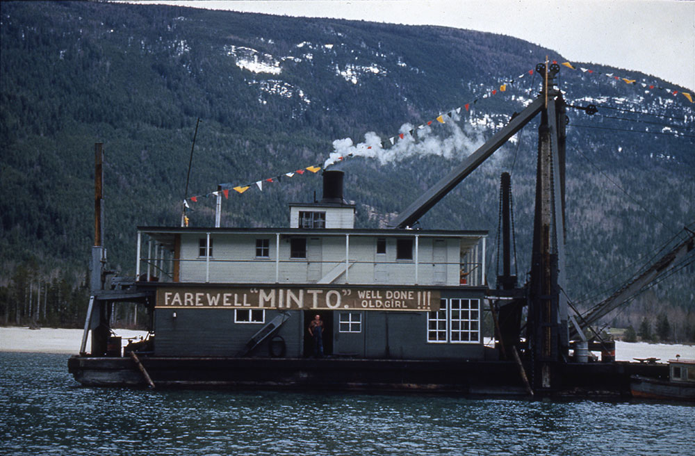 Une drague sur l’eau affiche un panneau en bois sur lequel on peut lire : « Farewell “MINTO”. Well done old girl!!! (Adieu “MINTO”. Bien joué ma vieille!!!) ». Un homme se tient dans l’embrasure de la porte, sous le panneau. Une guirlande de fanions triangulaires colorés est attachée de la cheminée jusqu’à l’avant du bateau. Une montagne se trouve à l’arrière-plan.