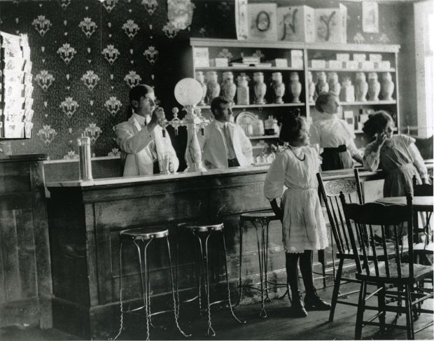 Photographie en noir et blanc à l’intérieur d’un bar laitier. Deux hommes et une femme se tiennent derrière un comptoir de bar sur lequel se trouve une machine à crème glacée. Des tabourets se trouvent devant le bar. Deux filles se tiennent devant le comptoir, derrière une table et des chaises. Le mur est recouvert de papier peint et devant se trouve une étagère remplie d’objets.