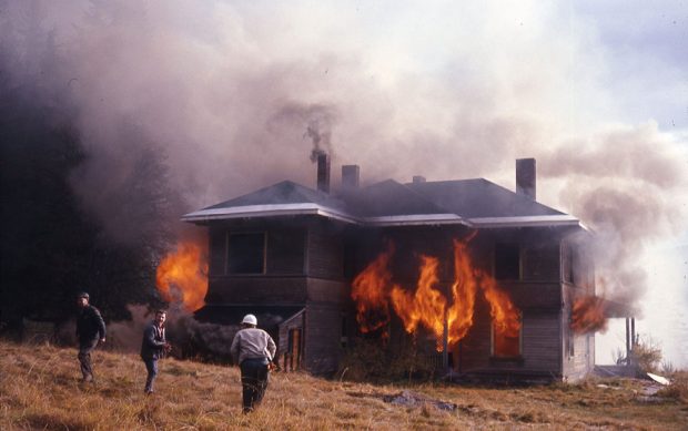 Photographie en couleur montrant trois personnes debout devant une maison en feu. La maison est encore intacte mais des flammes orange sortent des fenêtres. Une épaisse fumée blanche et noire entoure la maison.