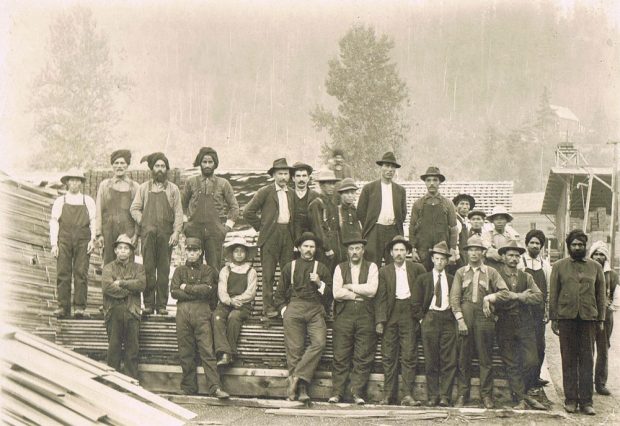 Photographie en noir et blanc d’un groupe d’hommes, une équipe de travail, debout devant un tas de bois. Ces hommes étaient d’origine japonaise, indienne, canadienne et européenne. Des arbres se trouvent en arrière-plan.