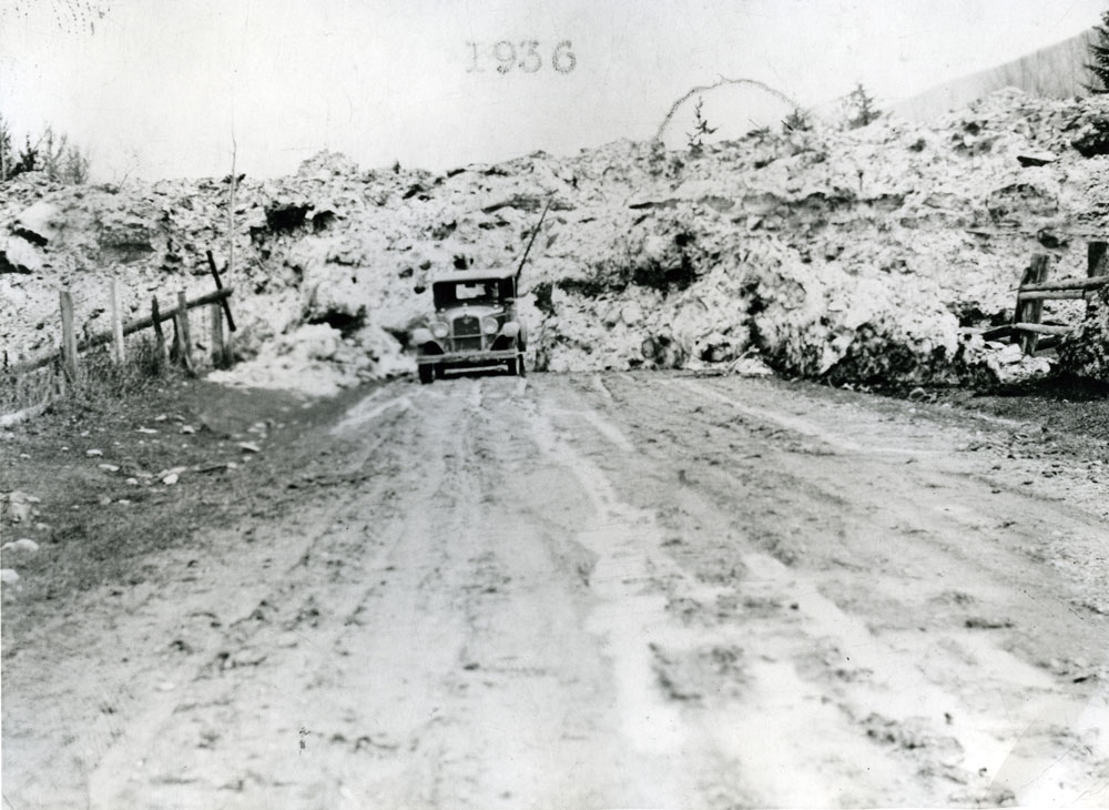 Photographie en noir et blanc d'une avalanche. Un immense banc de neige est en arrière-plan. Une voiture se trouve devant, sur la route. Il y a une clôture de chaque côté de la route.