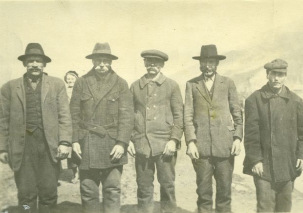 Photographie en échelle de gris de cinq hommes moustachus, debout, un à côté de l’autre. Les hommes portent des manteaux et des chapeaux. Une femme se trouve derrière le groupe, à gauche.