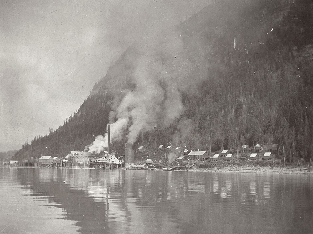 Photographie en noir et blanc d’une scierie située sur le bord d’une étendue d’eau et devant une montagne. Un nuage de fumée se dégage de la scierie. Il y a des maisons à côté de la scierie.