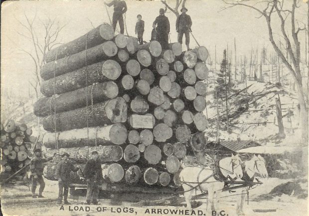 Quatre personnes sont debout sur une haute pile de plus de 50 grumes qui reposent sur une palette en bois attelée à deux chevaux blancs. Trois hommes sont au sol, à gauche du chargement de grumes. Une zone défrichée enneigée se trouve en arrière-plan. Au bas de la photographie, on peut lire « A load of logs, Arrowhead, B.C. (Chargement de grumes, Arrowhead, C.-B.) ».