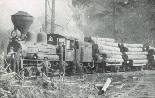 Photographie en noir et blanc d’un train transportant des grumes. Six hommes se tiennent devant le train, incluant le chef de train qui sort la tête par la fenêtre. Il y a des arbres en arrière-plan.