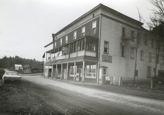 Photographie en noir et blanc d’un bâtiment à deux étages situé du côté droit d’un chemin de terre. Le haut du devant de l’édifice porte l’inscription « Union Hotel » et on y trouve deux enseignes à l’avant, l’une indiquant « Red & White food store » et l’autre « Temricks Store ». Il y a une voiture de l’autre côté de la rue, et des bâtiments et des arbres un peu plus loin.