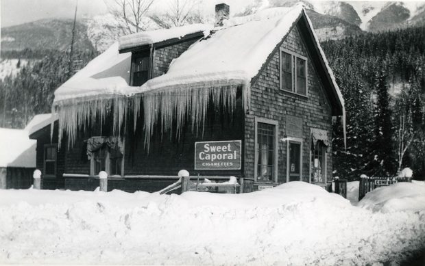 Photographie en noir et blanc d’un bâtiment enneigé avec de longs glaçons qui pendent du bord du toit. Une enseigne indiquant « Sweet Caporal Cigarettes » se trouve à la gauche du bâtiment. Le magasin est entouré d’une épaisse couche de neige. Des arbres enneigés et le pied d’une montagne se trouvent en arrière-plan.