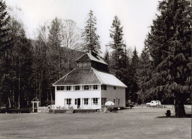 Photographie en noir et blanc d’un club house à deux étages situé dans un champ. Le patio du bâtiment est équipé de chaises d'extérieur. Une femme est assise sous un parasol à côté du bâtiment. Il y a une clôture à gauche du bâtiment. Des arbres et des voitures sur la route sont en arrière-plan.