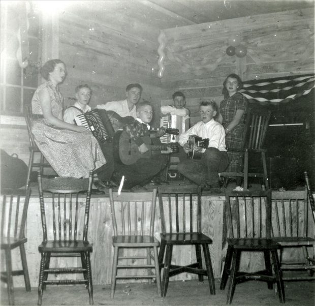 Photographie en noir et blanc de jeunes adolescents assis sur une scène, tenant des instruments de musique. Trois garçons tiennent des guitares et deux tiennent des accordéons. Deux filles se trouvent de chaque côté. Il y a des banderoles et des ballons accrochés au mur derrière la scène, et une rangée de chaises, en bas, devant la scène.