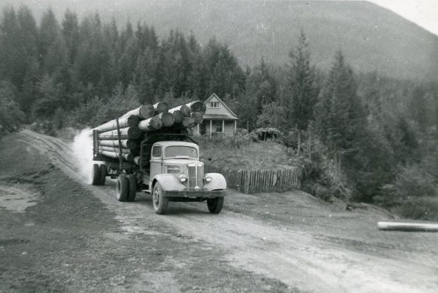 Photographie en noir et blanc d’un camion transportant des grumes sur un chemin de terre. Une maison se trouve derrière le camion. Des arbres et la base d’une montagne se trouvent à l’arrière-plan.