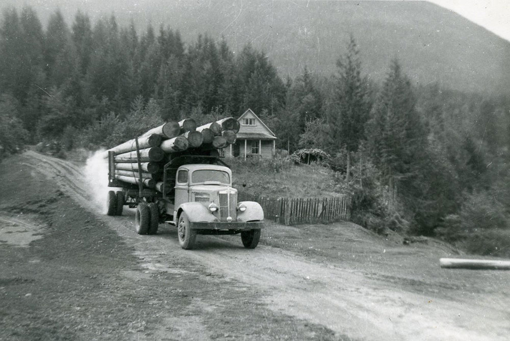 Photographie en noir et blanc d’un camion transportant des grumes sur un chemin de terre. Une maison se trouve derrière le camion. Des arbres et la base d’une montagne se trouvent à l’arrière-plan.