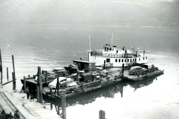 Photographie en noir et blanc d’une barge transportant une cargaison, dont quatre véhicules. La barge repose sur l'eau et une partie du quai est visible à gauche. Quatre personnes et deux chiens se tiennent sur le quai.