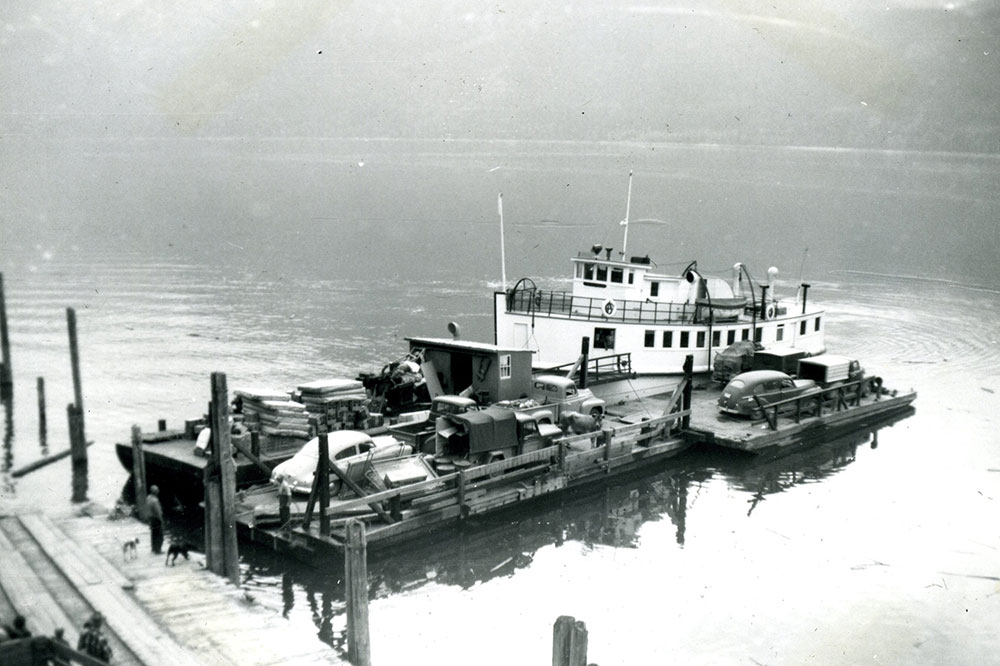 Photographie en noir et blanc d’une barge transportant une cargaison, dont quatre véhicules. La barge repose sur l'eau et une partie du quai est visible à gauche. Quatre personnes et deux chiens se tiennent sur le quai.