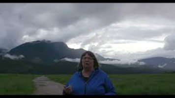 Cathy English – une femme blanche avec des cheveux courts gris, vêtue d’un manteau bleu avec une fermeture éclair. Elle se trouve sur un chemin de terre. Il s’y trouve de lʼherbe verte, une montagne et un ciel gris derrière elle.