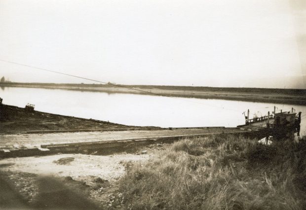 Photographie aux tons sépia d'un traversier amarré au bord de l'eau, avec une rampe d'embarquement menant au traversier.