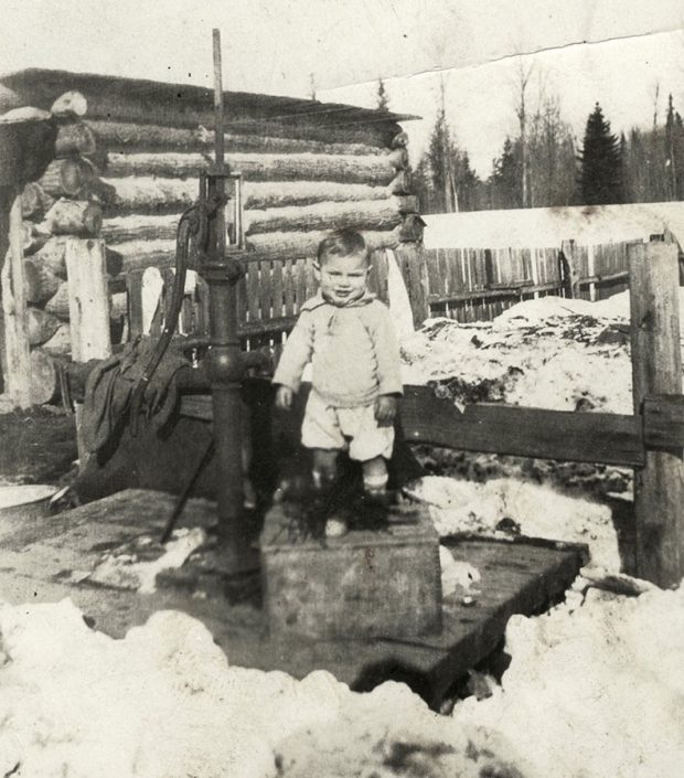 Photographie en noir et blanc d’un petit garçon debout sur une caisse en bois à côté d’une pompe à eau. Il y a une clôture derrière lui et un bâtiment en bois rond à sa gauche. C'est une journée d'hiver; on peut distinguer des tas de neige sale au sol.