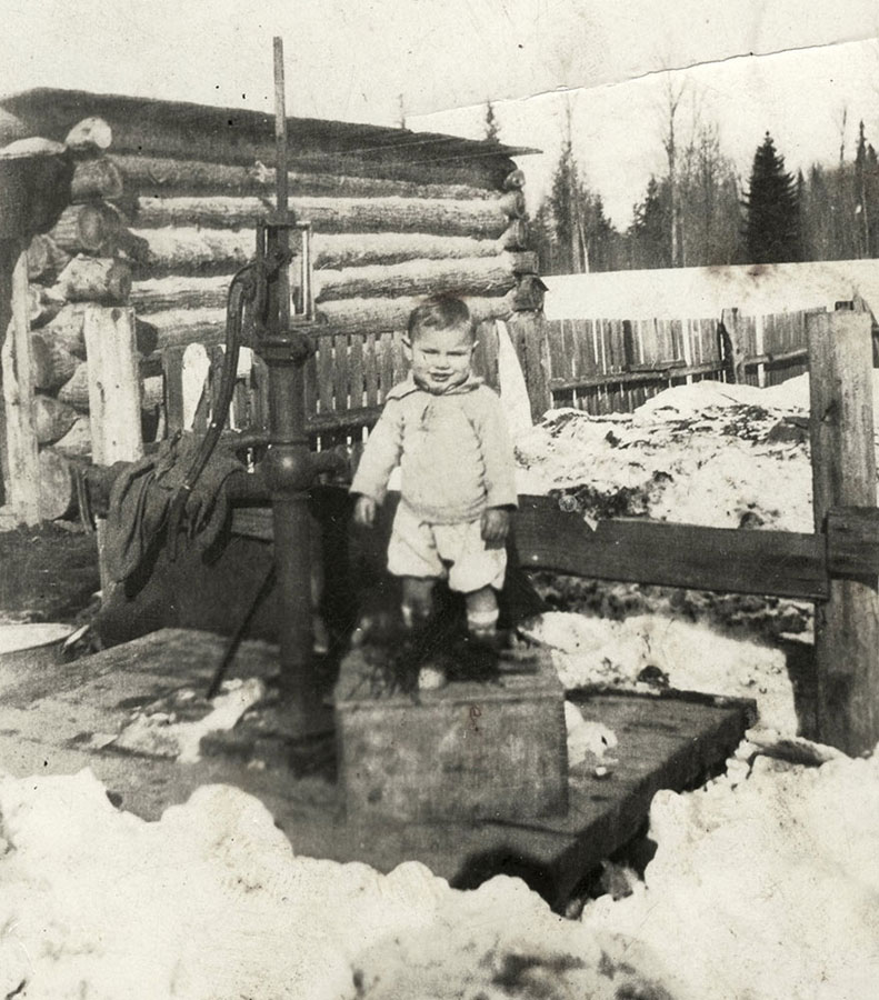 Photographie en noir et blanc d’un petit garçon debout sur une caisse en bois à côté d’une pompe à eau. Il y a une clôture derrière lui et un bâtiment en bois rond à sa gauche. C'est une journée d'hiver; on peut distinguer des tas de neige sale au sol.