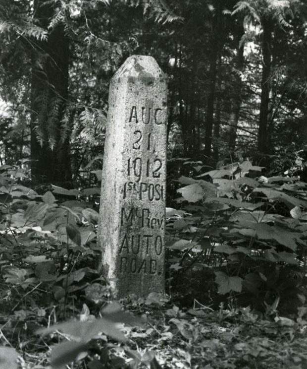 Photographie en noir et blanc d’un poteau de ciment qui a la forme d’un obélisque égyptien. On peut y lire « Aug 21 1912 1st Post Mt. Rev. Auto Road (21 août 1912 1er poteau route automobile du mont Revelstoke) ». Il est entouré d’arbres et de feuillage.