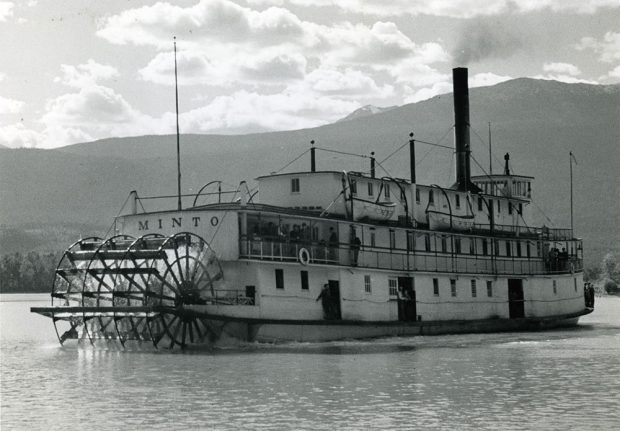 Photographie en noir et blanc d’un bateau à vapeur qui avance sur l’eau. La photo montre clairement la roue à aubes à la poupe du bateau, ainsi que le nom « MINTO » écrit juste au-dessus. Des montagnes sous un ciel partiellement nuageux se trouvent en arrière-plan.