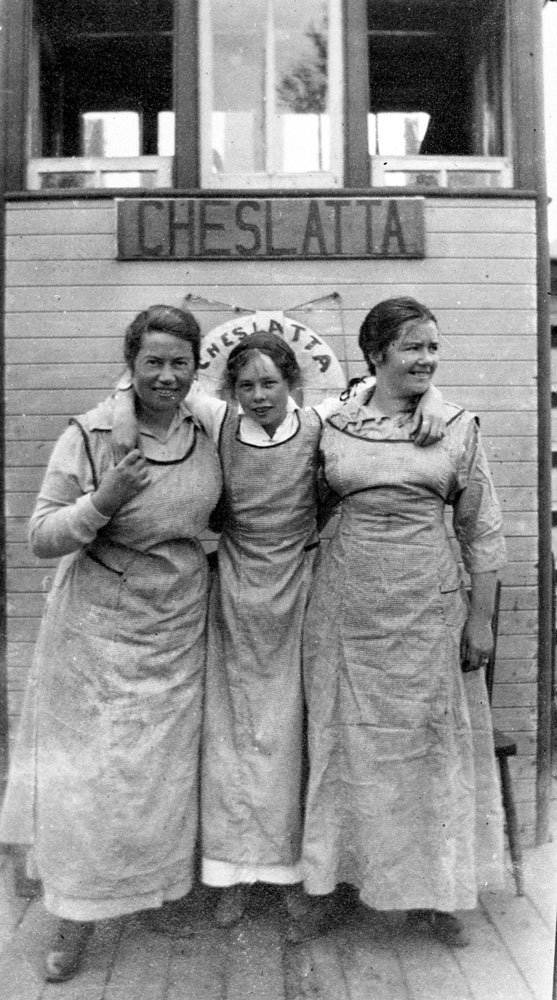 Photographie en noir et blanc de trois femmes debout ensemble. La femme au centre a ses bras sur les épaules des autres femmes. Elles se tiennent devant une bouée et un panneau sur lesquels on peut lire « Cheslatta ».