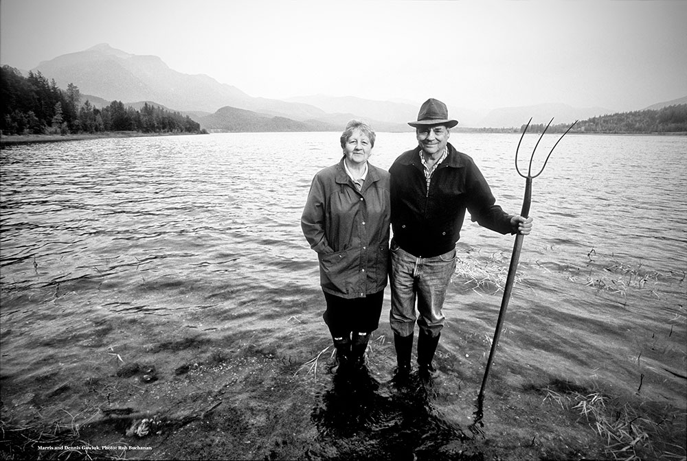 Photographie en noir et blanc d’une femme et d’un homme se tenant debout dans une rivière, avec de l’eau jusqu’aux chevilles. L’homme tient une fourche. Il y a des montagnes et des arbres en arrière-plan.