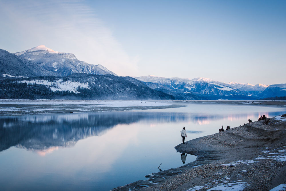 Photographie en couleur de montagnes enneigées se reflétant sur une étendue d'eau. Une personne se tient de dos sur la berge rocailleuse.