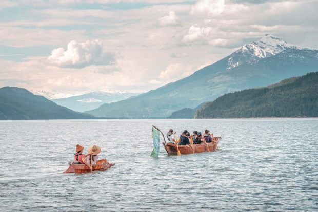 Photographie en couleur de deux personnes dans un canot à nez d’esturgeon pagayant derrière un grand canot creusé, arborant le drapeau Sinixt. Sous un ciel partiellement nuageux, les bateaux avancent sur une rivière, avec des montagnes en arrière-plan.