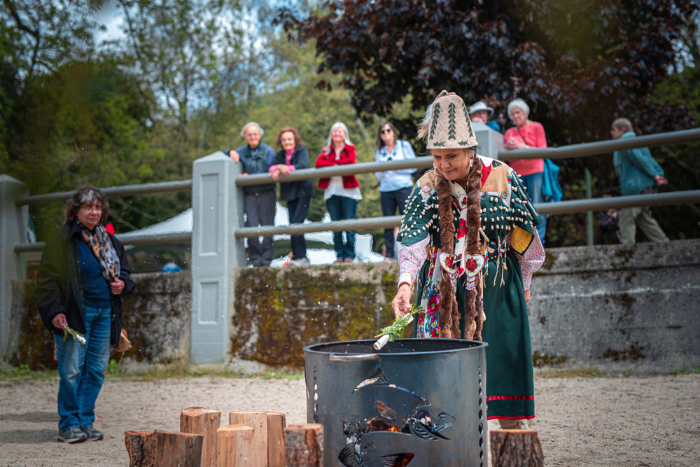 Photographie en couleur d’une femme Sinixt vêtue d’une tenue traditionnelle, jetant un document enroulé dans un baril spécialement construit pour la combustion. Une personne tenant un document similaire se tient un peu plus loin, à gauche. En arrière-plan, il y a des gens flous qui observent derrière un garde-corps sur une terrasse surélevée. Des arbres sont aussi en arrière-plan. 