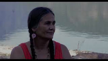 Shelly Boyd – une femme Sinixt avec deux longues tresses noires, vêtue d'un débardeur orange. Ceci est un portrait de Shelly avec de l’eau derrière elle.