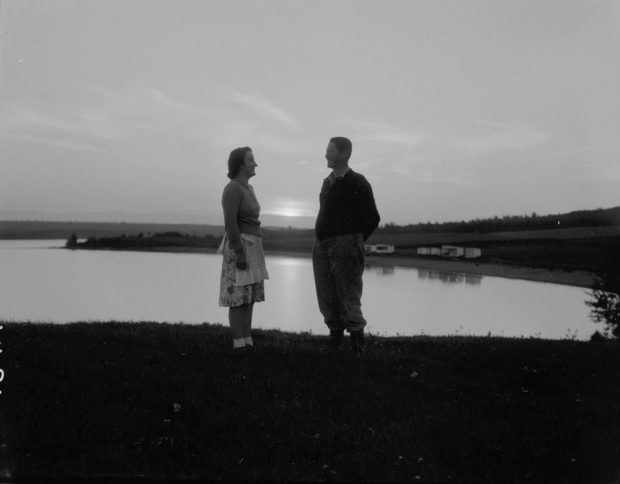 Photographie noir et blanc d'un couple debout, se faisant face, devant un lac, au soleil couchant.