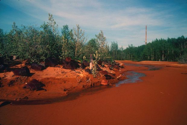 Photographie couleur d'un cours d'eau polluée, d'une teinte très orangée : des barils de métal rouillés traînent dans la forêt. À l'arrière-plan, on distingue une très haute cheminée d'une fonderie.