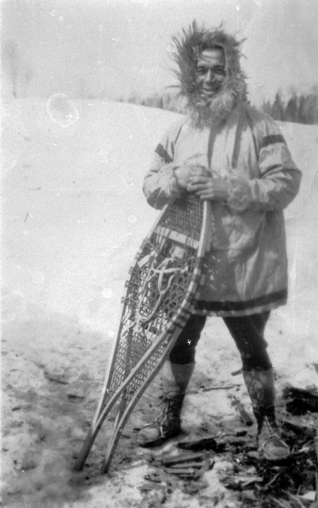 Photographie noir et blanc d'un homme en parka qui tient une paire de raquettes, en hiver, à l’extérieur.