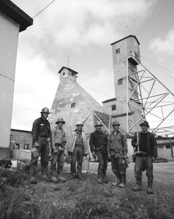 Six mineurs en habit de travail posent debout devant l'échafaud d'une mine et d'autres édifices industriels.