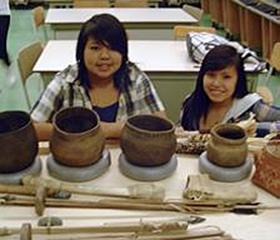 Des reproductions de vases et autres objets de culture algonquine disposés sur une table. Deux adolescents regardent le photographe, en arrière-plan.