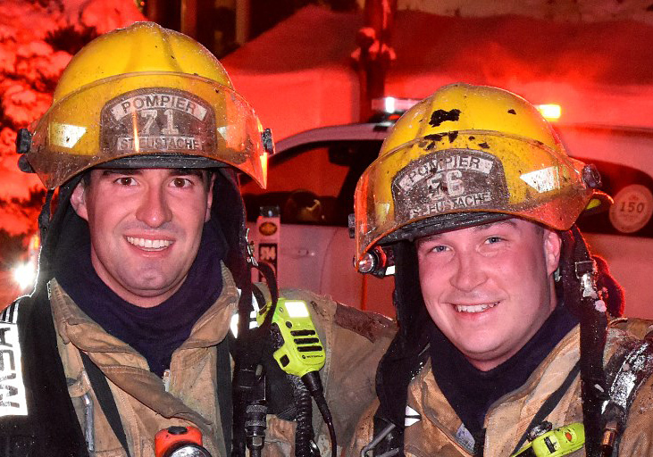 Photographie de deux pompiers souriants en tenue de combat contre le feu, devant un camion de pompiers, une voiture et plusieurs maisons lors d’une nuit éclairée.
