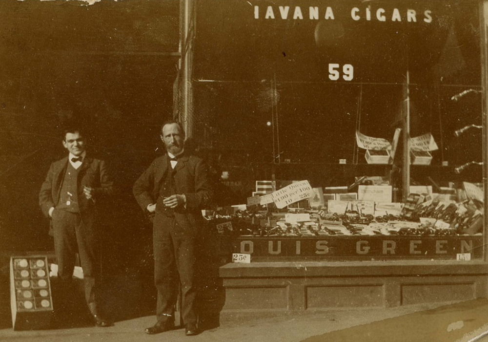 Devanture dotée d’une grande vitrine. En haut, on peut lire « Havana Cigars » et en bas, « Louis Green ». La vitrine expose des cigares et des produits de tabac. Deux hommes se tiennent debout dans l’entrée.