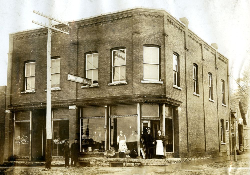 Photo en noir et blanc datant d’environ 1914 d’un magasin d’aliments secs à la façade en brique. Trois hommes se tiennent à l’intérieur du magasin et quatre se tiennent à l’extérieur, tandis que l’enseigne « Dominion Hall » surplombe la rue. La devanture de l’établissement de gauche porte l’inscription « E.H. Cottrell Meat Market » (Halles aux viandes E.H. Cottrell). La rue de terre est humide et boueuse.