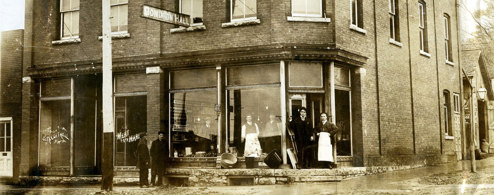 Photo en noir et blanc datant d’environ 1914 d’un magasin d’aliments secs à la façade en brique. Trois hommes se tiennent à l’intérieur du magasin et quatre se tiennent à l’extérieur, tandis que l’enseigne « Dominion Hall » surplombe la rue. La devanture de l’établissement de gauche porte l’inscription « E.H. Cottrell Meat Market » (Halles aux viandes E.H. Cottrell). La rue de terre est humide et boueuse.