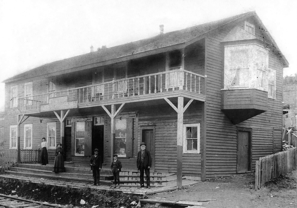 Photo en noir et blanc du British North American Hotel de la famille Joseph, vers le début des années 1900, sur laquelle on peut voir un grand bâtiment de deux étages. Deux femmes, deux jeunes garçons et un homme plus âgé se tiennent debout sur les marches du bâtiment. Il y a une clôture autour du bâtiment et, devant les marches, il y a des rails de chemin de fer.