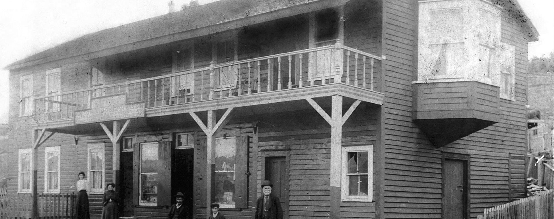 Photo en noir et blanc du British North American Hotel de la famille Joseph, vers le début des années 1900, sur laquelle on peut voir un grand bâtiment de deux étages. Deux femmes, deux jeunes garçons et un homme plus âgé se tiennent debout sur les marches du bâtiment. Il y a une clôture autour du bâtiment et, devant les marches, il y a des rails de chemin de fer.