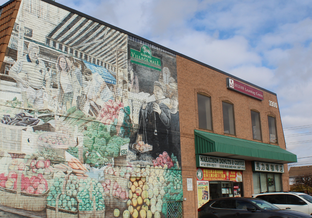 Une photographie couleur d'un bâtiment en briques rouges de deux étages avec des fenêtres cintrées, des devantures de magasins et une grande fresque peinte à la main sur la moitié du bâtiment représentant un marché de producteurs.